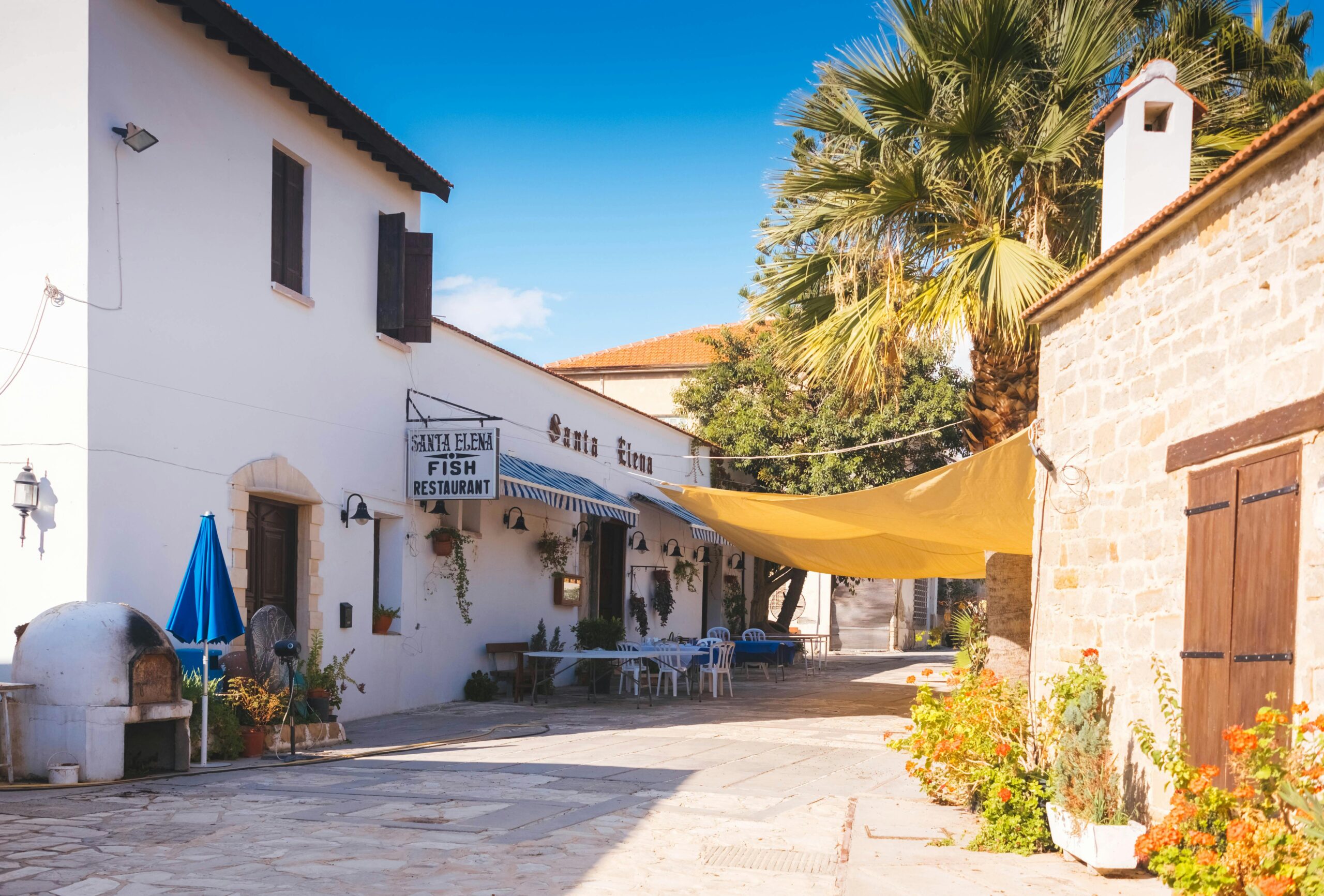 Exterior view of a quaint restaurant in Cyprus with outdoor seating, palm trees, and stone architecture.